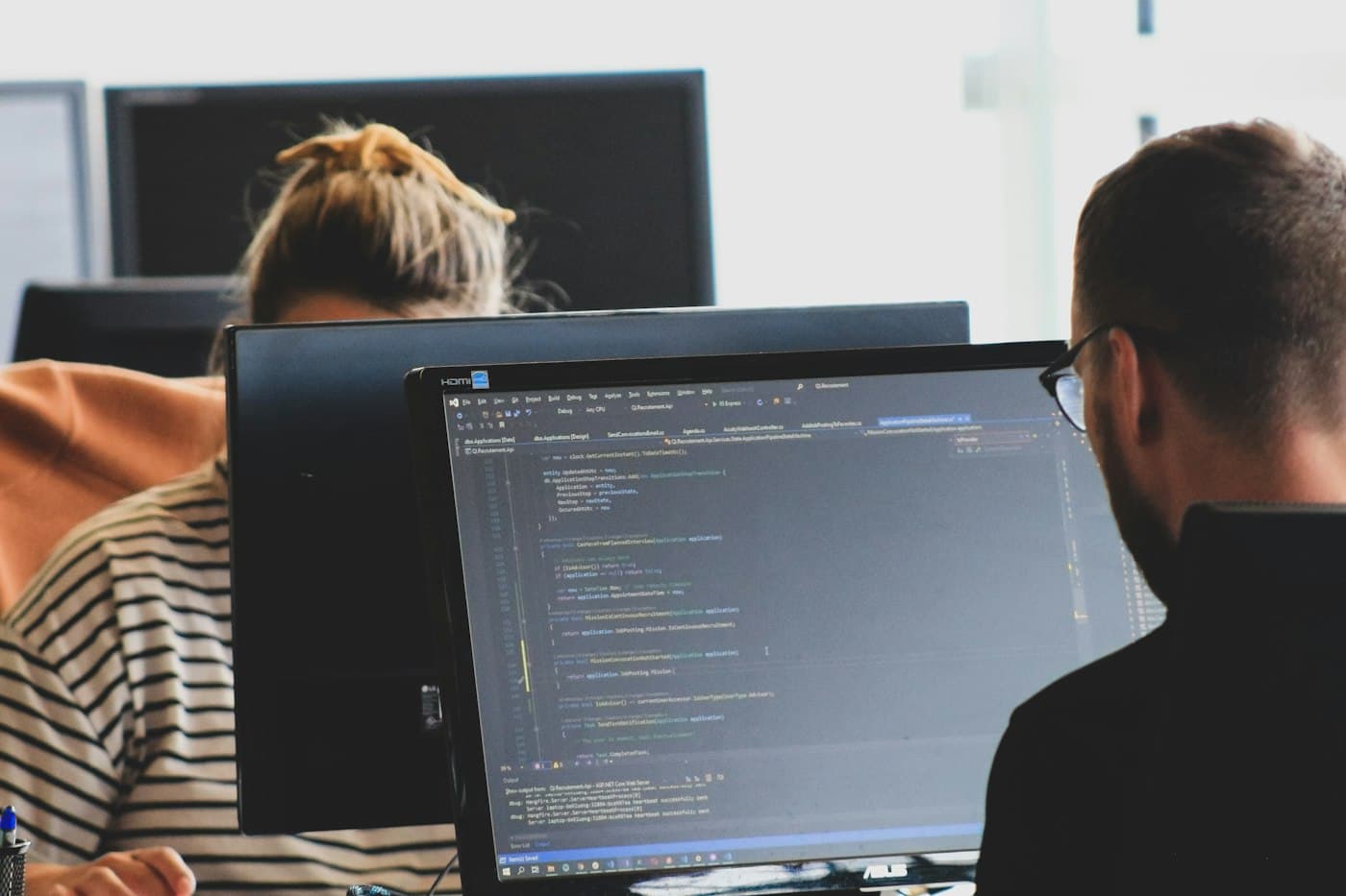 Dos personas trabajando de espaldas frente a monitores con código en pantalla